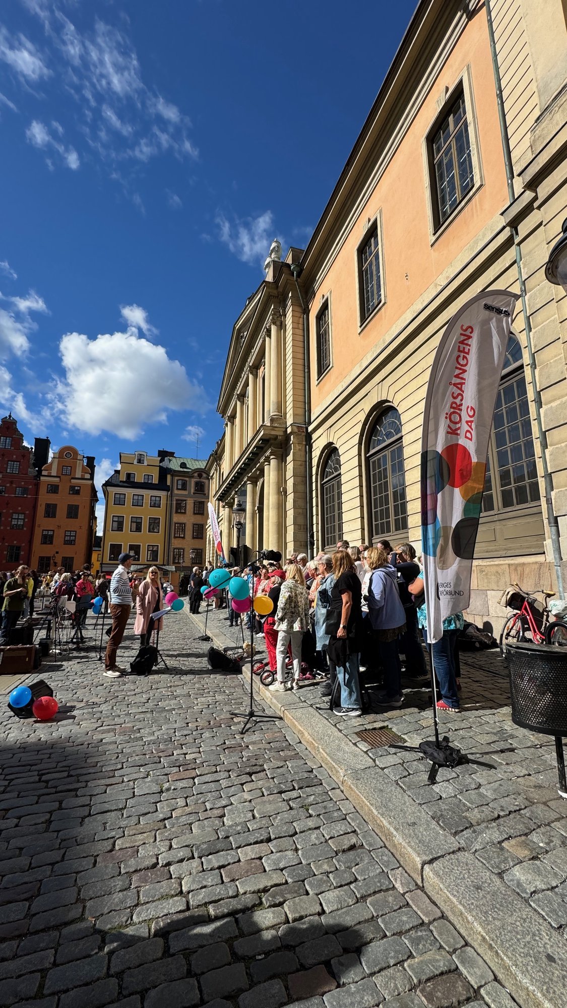 A choir and a crowd with balloons on the cobblestones of Stortorget, beside the yellow Börshuset building. Colored merchant houses stand behind.
