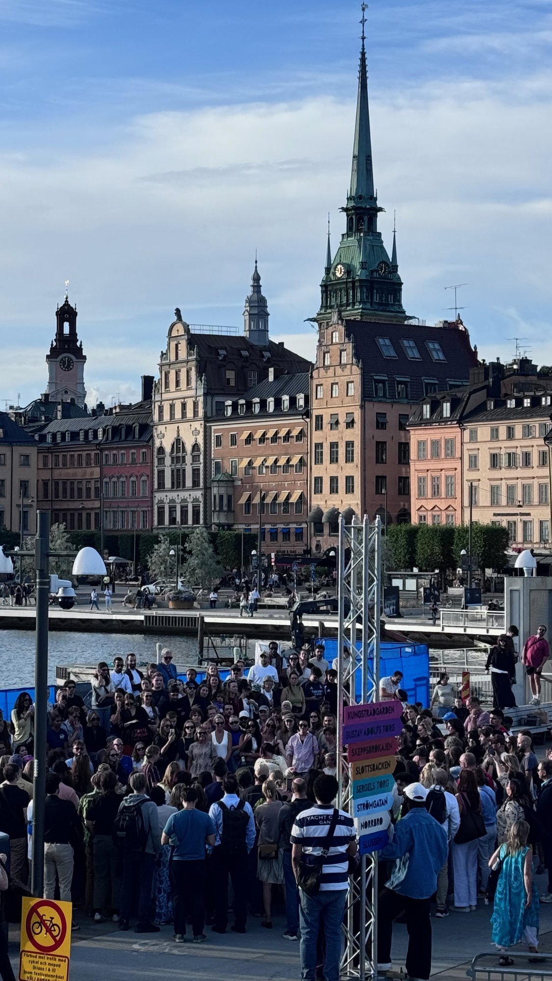 A large crowd stands in a festival area in summer. Behind it rise the old houses of Gamla Stan and the green spire of Tyska kyrkan.