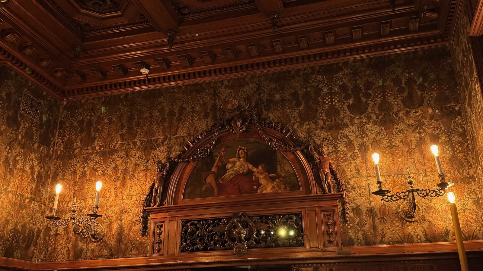 A carved wooden wall panel with a painting, gold patterned walls and candle lights at the Hallwyl Museum.