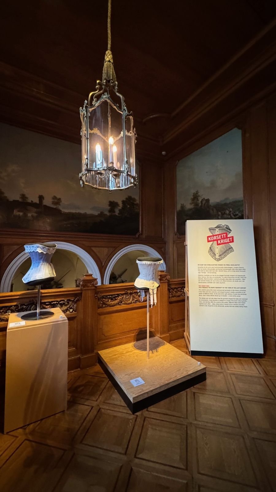 White corsets on stands under a hanging lantern, next to the Korsettkriget sign, at the Hallwyl Museum.