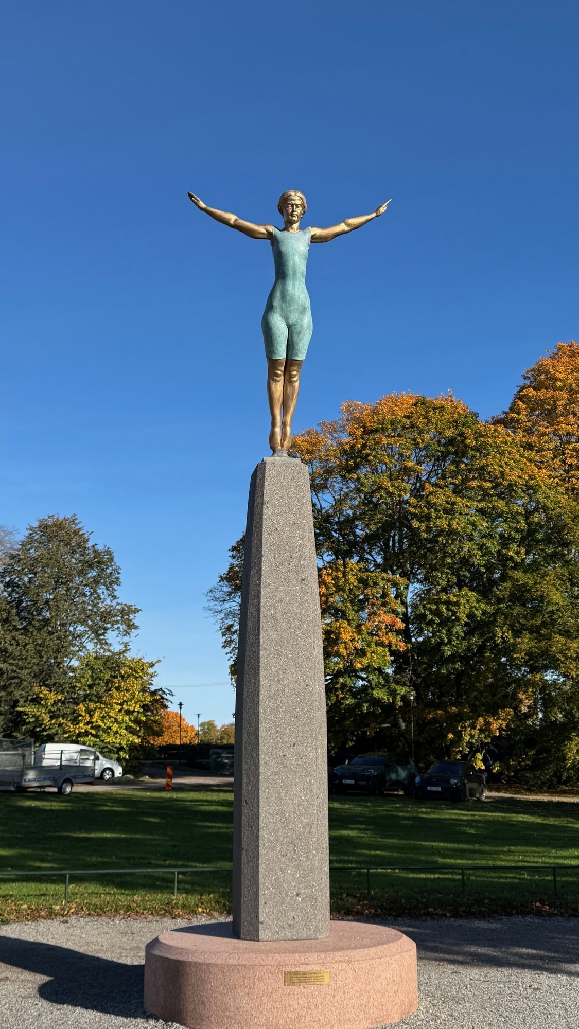 Bronze statue of diver Greta Johansson with her arms spread wide, on a tall stone column, with autumn trees behind.