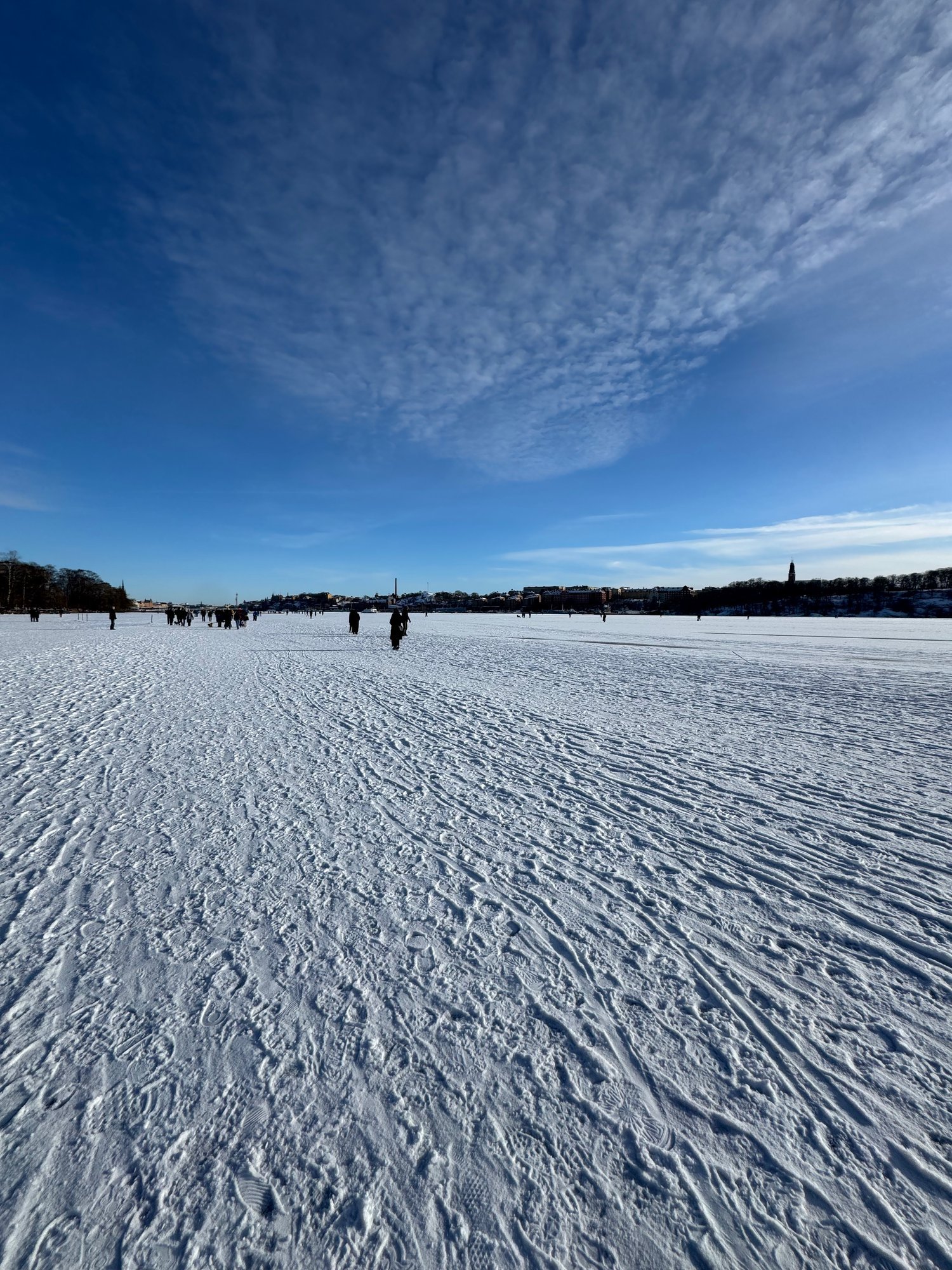 Wind has drawn wavy lines in the snow on frozen Riddarfjärden. Small figures of people walk on the ice far away, under a blue sky.