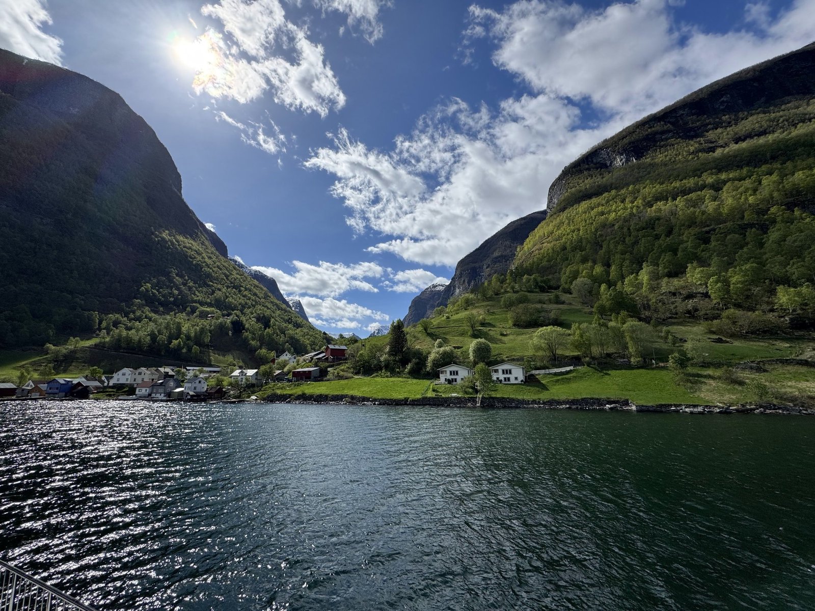 A village on the green shore of the Nærøyfjord, below steep mountains, on a sunny day.