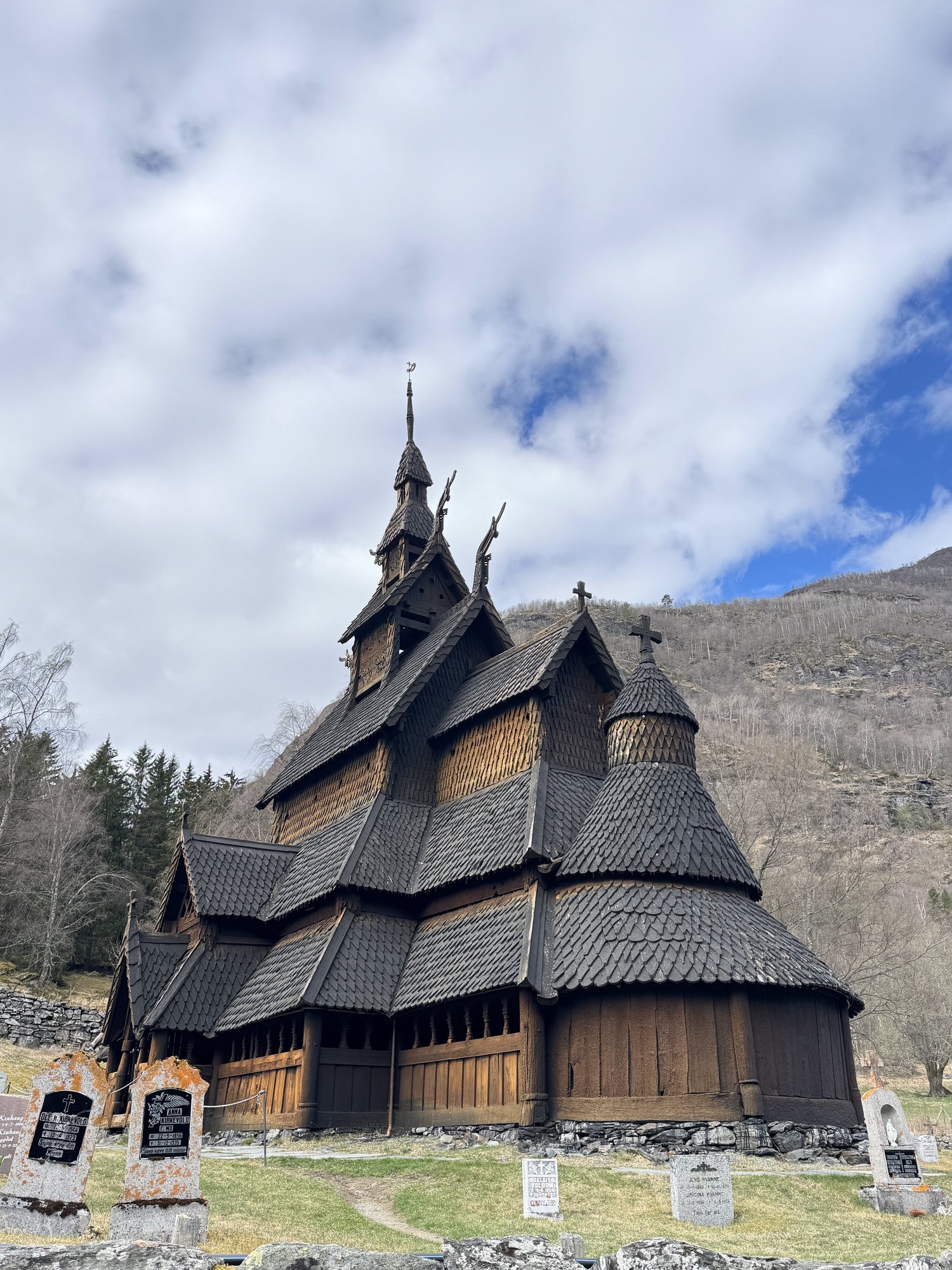 A dark wooden stave church with many roofs and carved dragon heads, in a churchyard.