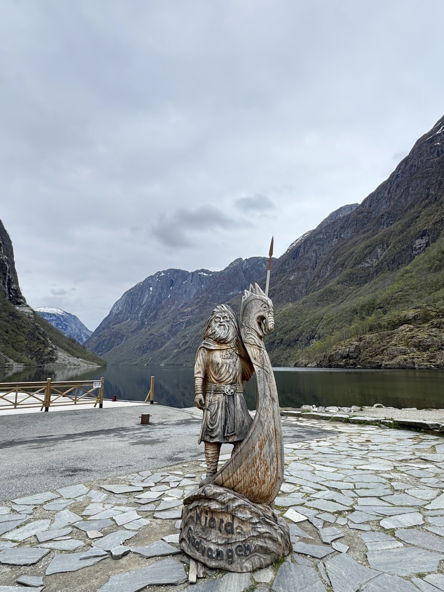 A carved wooden Viking statue with a dragon-head prow, on a stone square by a fjord.