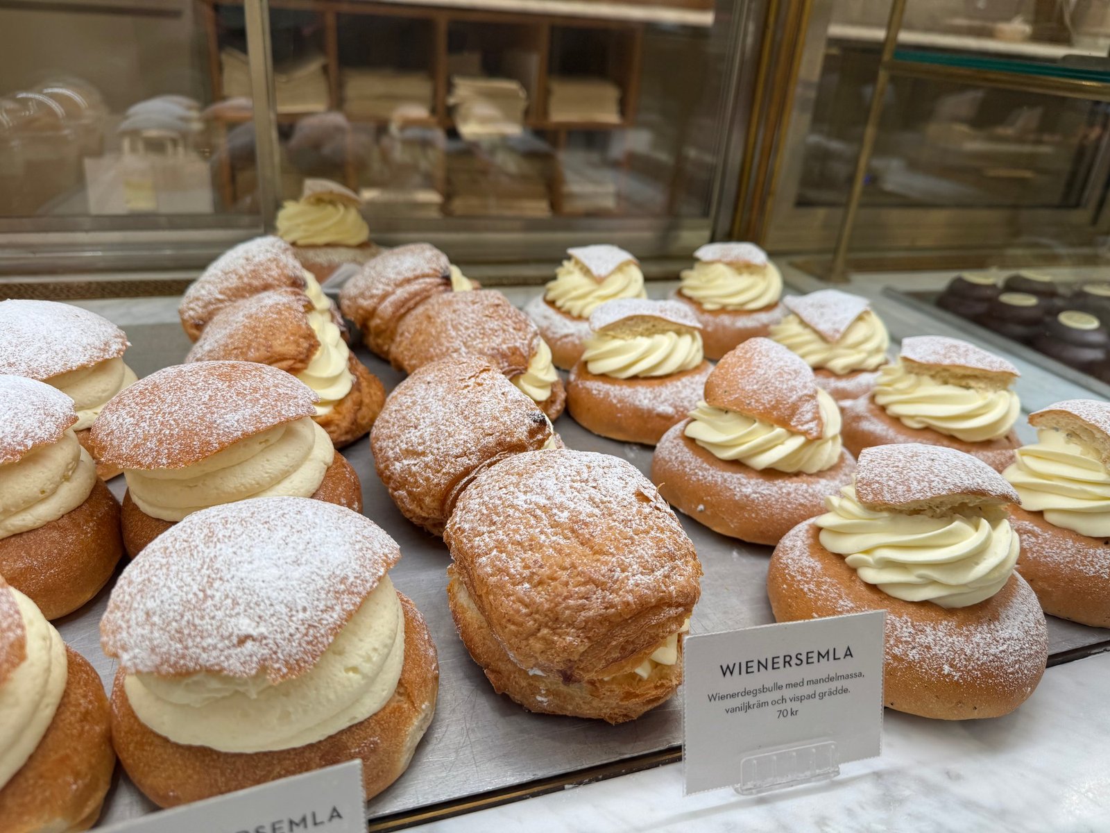 Semla buns filled with whipped cream and dusted with powdered sugar, on a tray in a bakery display.