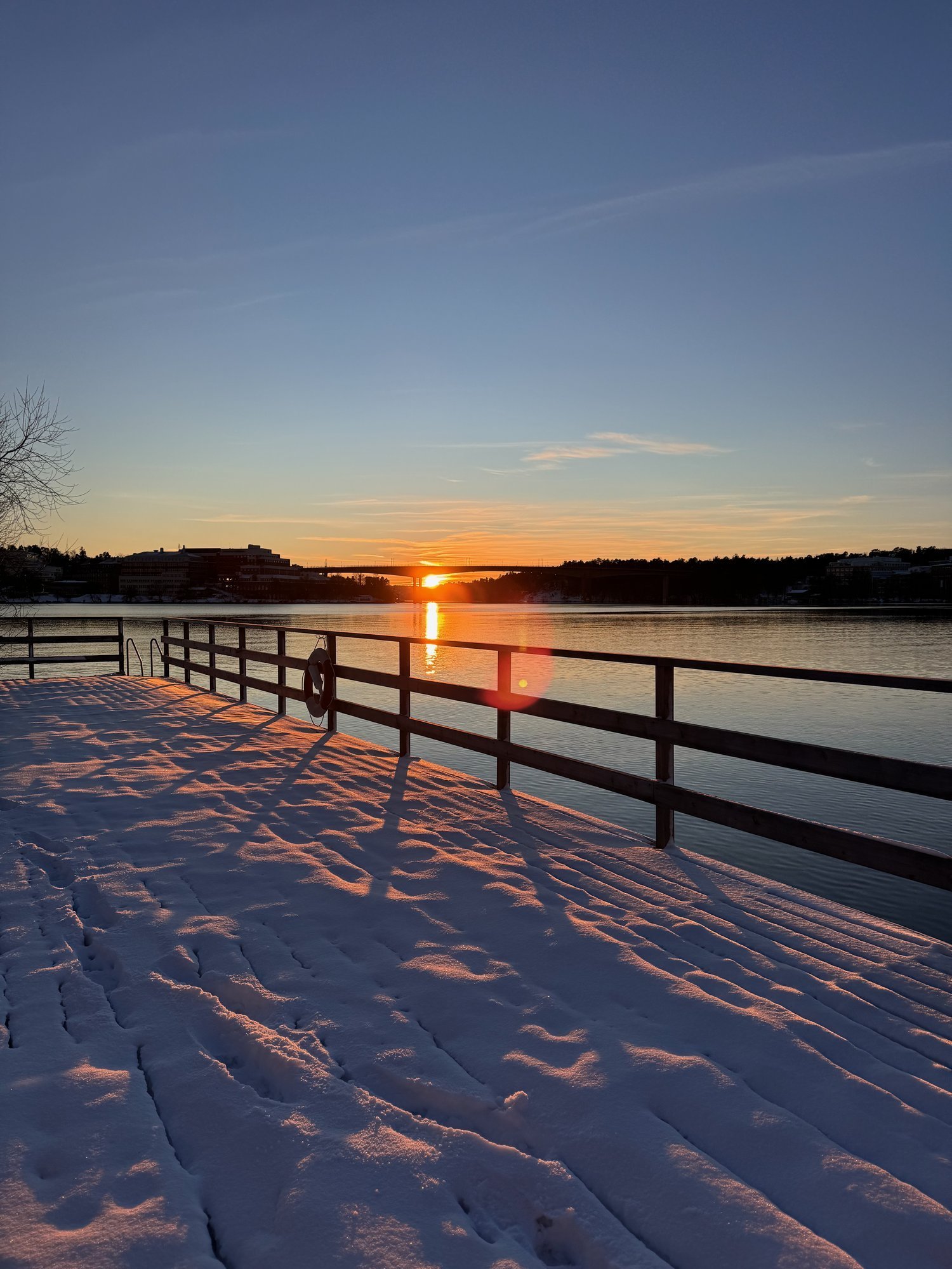 A snowy wooden jetty with a railing runs along a lake at sunset.