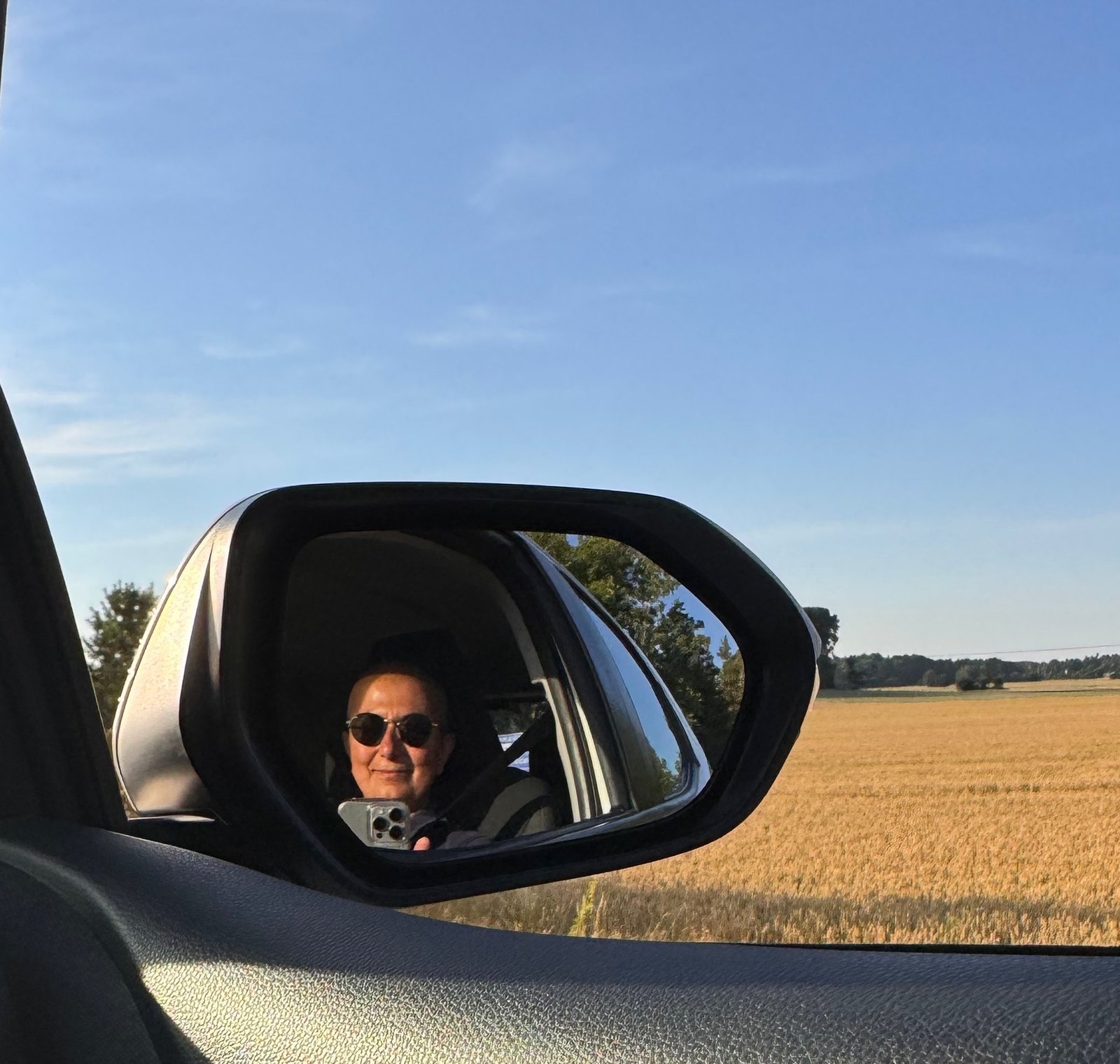 Zayera Khan seen in the car mirror on a road trip past a wheat field in Sweden